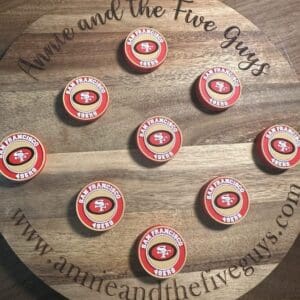 Round wooden board with "Annie and the Five Guys" and website text, holding eight red and white San Francisco football silicone focal beads.