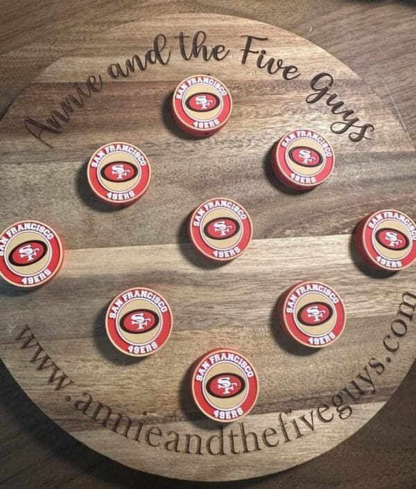 Round wooden board with "Annie and the Five Guys" and website text, holding eight red and white San Francisco football silicone focal beads.