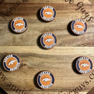 Seven Denver football silicone focal beads are displayed on a round wooden board with the text "Annie and the Five Guys, www.annieandthefiveguys.com" engraved on it.