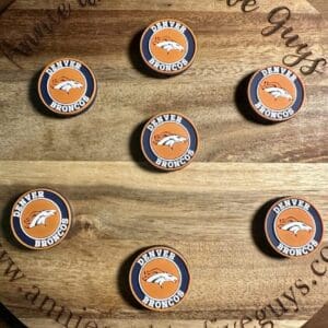 Seven Denver football silicone focal beads are displayed on a round wooden board with the text "Annie and the Five Guys, www.annieandthefiveguys.com" engraved on it.