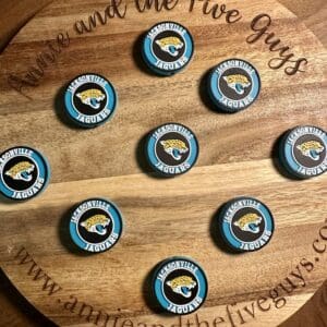 A display featuring nine Jacksonville football silicone focal beads arranged in a circular pattern is set on a wooden board with engraved text. The board reads "Amie and the Five Guys" and includes a website URL on the edge.