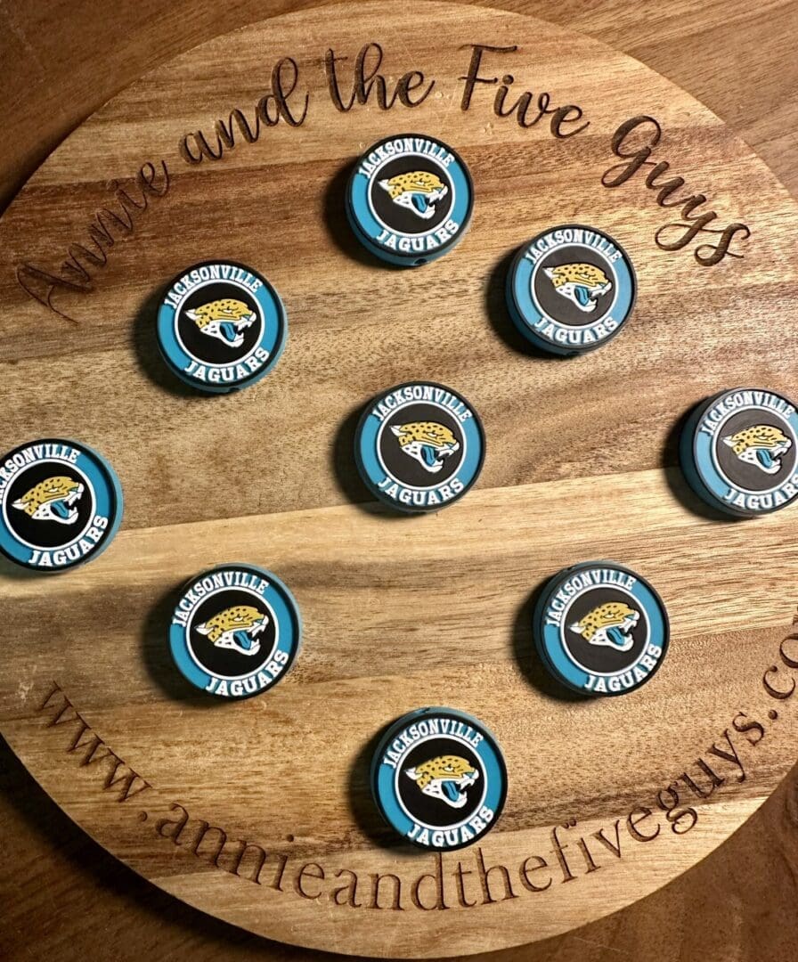 A display featuring nine Jacksonville football silicone focal beads arranged in a circular pattern is set on a wooden board with engraved text. The board reads "Amie and the Five Guys" and includes a website URL on the edge.