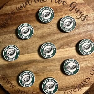 A wooden board with "www.annieandthefiveguys.com" engraved on it displays nine Philadelphia Eagles logo pins arranged in a circular pattern.