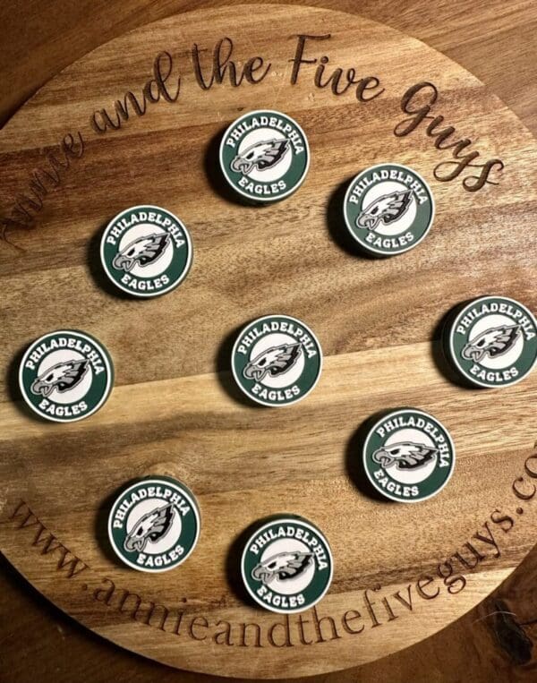 A wooden board with "www.annieandthefiveguys.com" engraved on it displays nine Philadelphia Eagles logo pins arranged in a circular pattern.