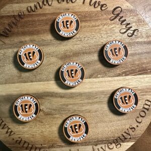 Seven circular silicone focal beads featuring the Cincinnati football logo are arranged on a wooden board inscribed with "Annie and the Five Guys" and a website URL.