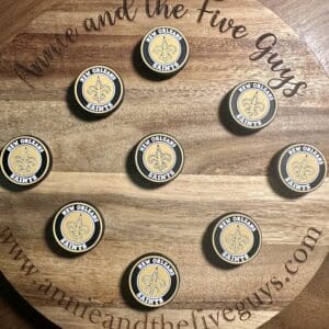 Round wooden board with nine New Orleans Saints logo magnets arranged on it. The board has the text "Annie and the Five Guys" and a website URL along the edges.