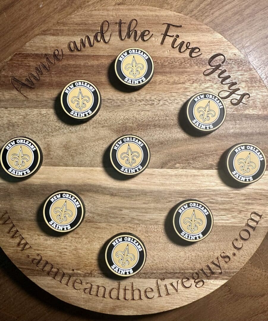 Round wooden board with nine New Orleans Saints logo magnets arranged on it. The board has the text "Annie and the Five Guys" and a website URL along the edges.