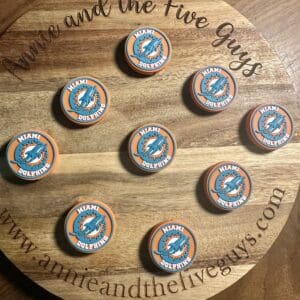 A wooden board displaying nine orange and teal Miami football silicone focal beads arranged in a semi-circular pattern. The board is branded with "Annie and the Five Guys" website text.