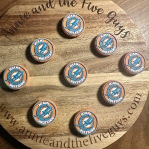 A wooden board displaying nine orange and teal Miami football silicone focal beads arranged in a semi-circular pattern. The board is branded with "Annie and the Five Guys" website text.