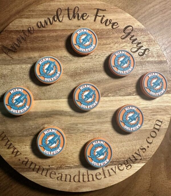 A wooden board displaying nine orange and teal Miami football silicone focal beads arranged in a semi-circular pattern. The board is branded with "Annie and the Five Guys" website text.