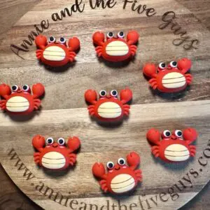 A wooden board features eight crab-themed macarons with red shells and animated eyes. The board is inscribed with "Annie and the Five Guys" and a website URL.