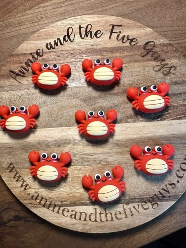 A wooden board features eight crab-themed macarons with red shells and animated eyes. The board is inscribed with "Annie and the Five Guys" and a website URL.