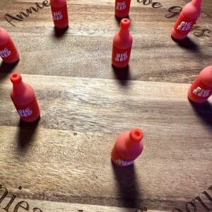 Seven small, red toy bottles labeled "Big Red" are arranged in a loose circle on a wooden surface. Text engraved on the wood reads "Anne and the Five Guys.