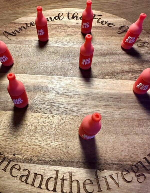 Seven small, red toy bottles labeled "Big Red" are arranged in a loose circle on a wooden surface. Text engraved on the wood reads "Anne and the Five Guys.