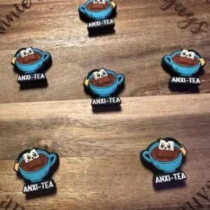 Several enamel pins shaped like teacups labeled "ANXI-TEA," featuring a stressed teabag character. They are displayed on a wooden background with the words "Annie and the Five Guys" engraved.