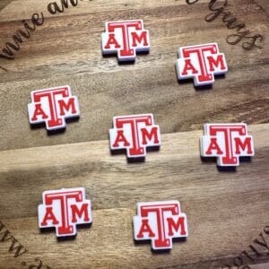 Wooden board featuring seven Texas A&M silicone focal beads in red and white. The board includes text "Annie and the Five Guys" along with "www.annieandthefiveguys.com" around its edges.