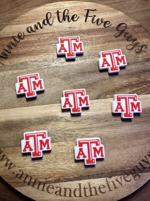 Wooden board featuring seven Texas A&M silicone focal beads in red and white. The board includes text "Annie and the Five Guys" along with "www.annieandthefiveguys.com" around its edges.