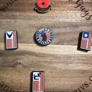 Five veteran-themed enamel pins on a wooden surface: a poppy, a circle with "Proud to be a Veteran," and three with U.S. flag designs and military symbols.