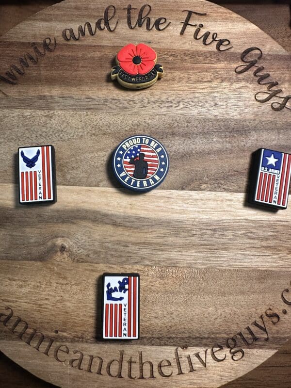 Five veteran-themed enamel pins on a wooden surface: a poppy, a circle with "Proud to be a Veteran," and three with U.S. flag designs and military symbols.