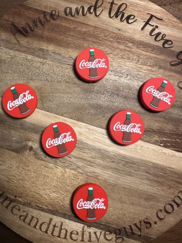 Six round, red Coca-Cola coasters with bottle designs are arranged on a wooden surface with partial website text.