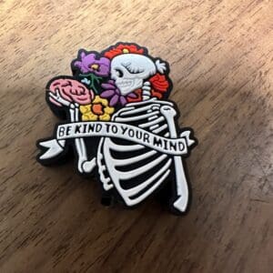 A skeleton pin holding a brain and flowers, with a banner reading "Be kind to your mind.