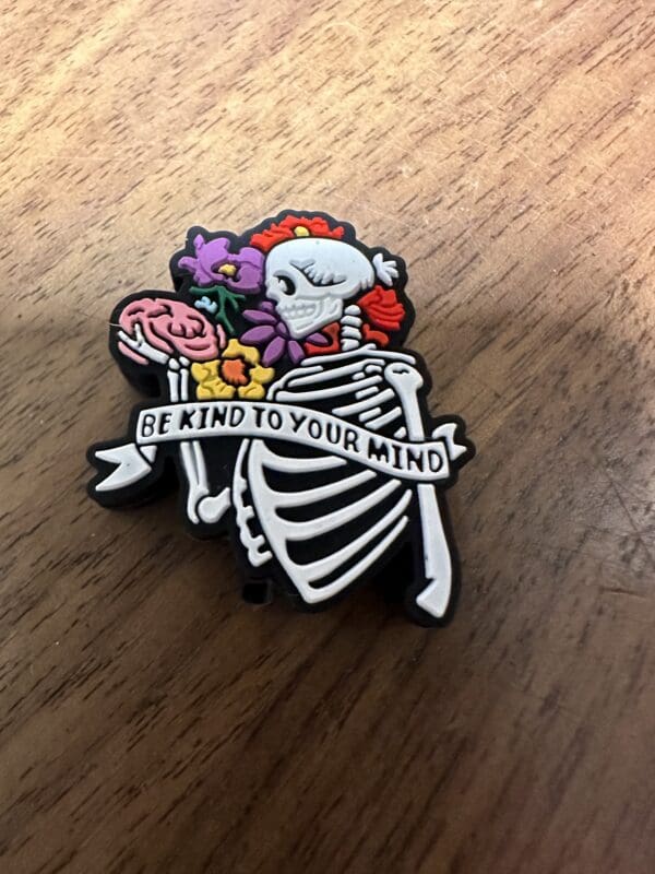 A skeleton pin holding a brain and flowers, with a banner reading "Be kind to your mind.