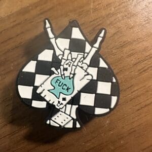 Pin with a spade shape, featuring a checkered pattern and a hand making a rock gesture. The hand holds a speech bubble with the word "FUCK.