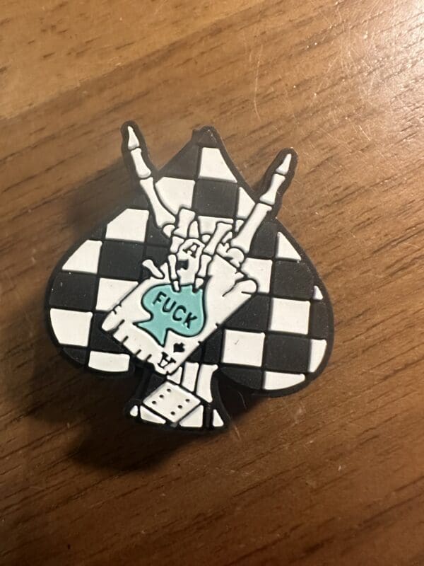 Pin with a spade shape, featuring a checkered pattern and a hand making a rock gesture. The hand holds a speech bubble with the word "FUCK.