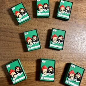 Nine green matchboxes with "Los Marijuanos" and cartoon characters on a wooden surface.