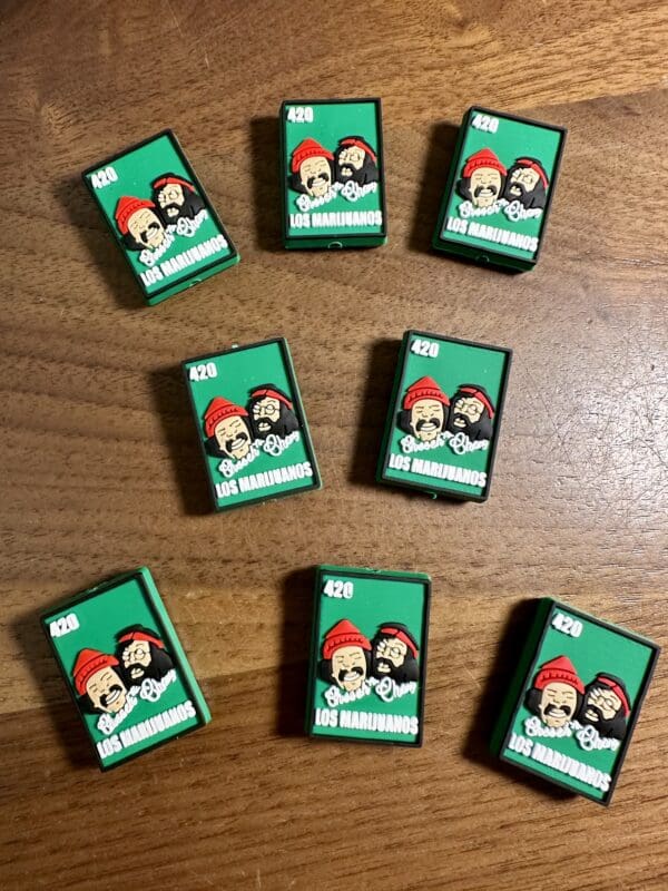 Nine green matchboxes with "Los Marijuanos" and cartoon characters on a wooden surface.