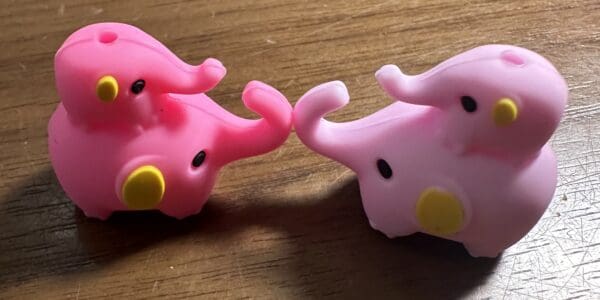 Two small, pink plastic elephant figurines with yellow accents face each other on a wooden surface.