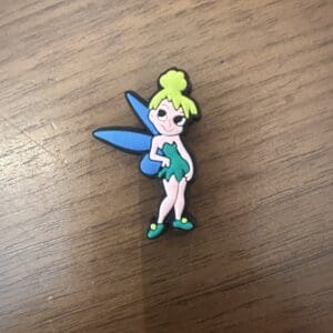 Pin depicting a cartoon fairy with green hair, blue wings, and a green dress, standing on a wooden surface.