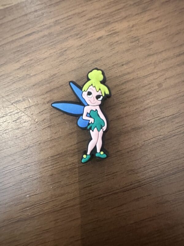 Pin depicting a cartoon fairy with green hair, blue wings, and a green dress, standing on a wooden surface.