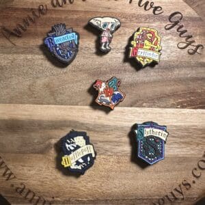 Six painted pins featuring various "Harry Potter" designs, including house crests and an elf, are displayed on a wooden board with engraved text.