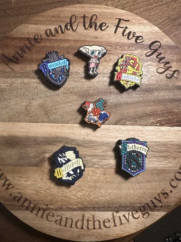 Six painted pins featuring various "Harry Potter" designs, including house crests and an elf, are displayed on a wooden board with engraved text.