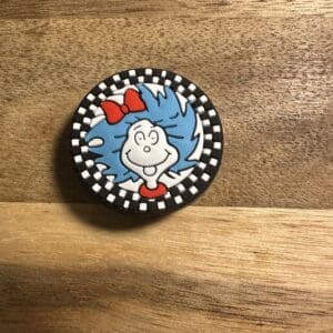 A round pin featuring an animated character with blue hair and a red bow, set against a black-and-white checkered border, placed on a wooden surface.