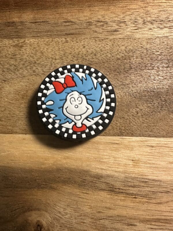 A round pin featuring an animated character with blue hair and a red bow, set against a black-and-white checkered border, placed on a wooden surface.