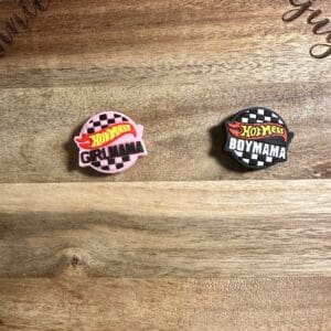 Two enamel pins on a wooden surface. One reads "Hot Mess Girl Mama" in pink, and the other reads "Hot Mess Boy Mama" in black. Both feature a checkered design.