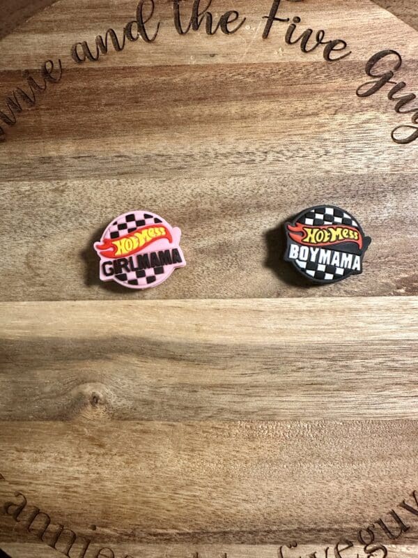 Two enamel pins on a wooden surface. One reads "Hot Mess Girl Mama" in pink, and the other reads "Hot Mess Boy Mama" in black. Both feature a checkered design.