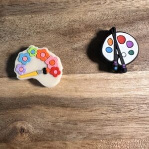 Two paint palette pins on a wooden surface. One pin has colorful flowers, the other has a classic palette with brushes.