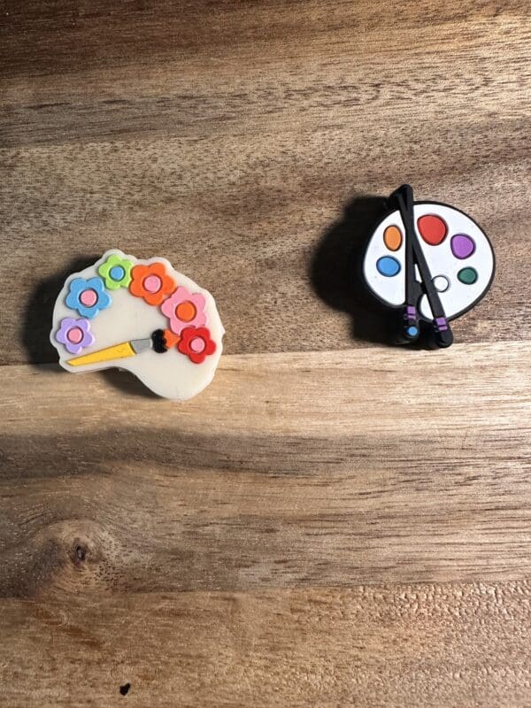 Two paint palette pins on a wooden surface. One pin has colorful flowers, the other has a classic palette with brushes.