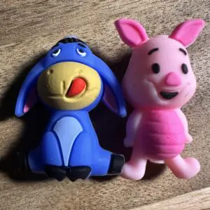 Two plastic toy figures on a wooden surface, one is a blue donkey with a red tongue sticking out, and the other is a pink pig with a smiling face.