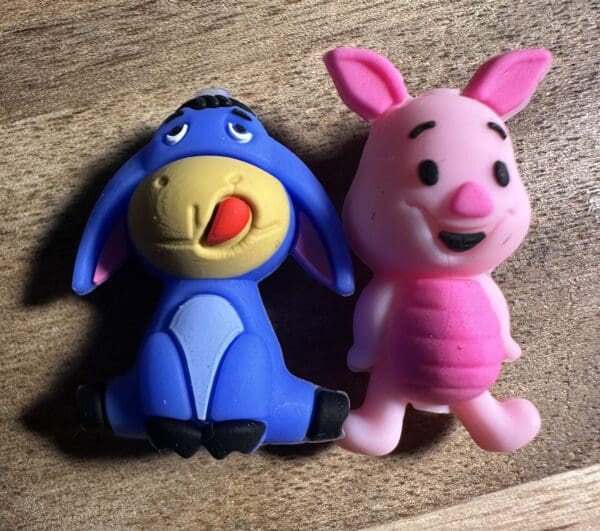 Two plastic toy figures on a wooden surface, one is a blue donkey with a red tongue sticking out, and the other is a pink pig with a smiling face.