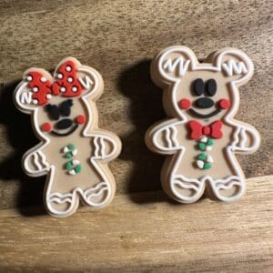 Two gingerbread bear cookies on a wooden surface. One wears a red bow with white polka dots, the other has a green bow tie. Both have decorated icing details.