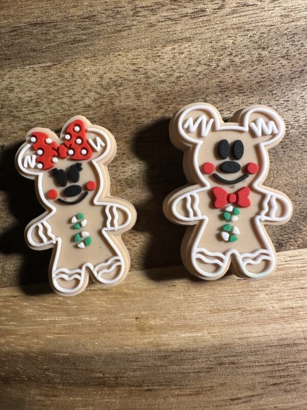 Two gingerbread bear cookies on a wooden surface. One wears a red bow with white polka dots, the other has a green bow tie. Both have decorated icing details.