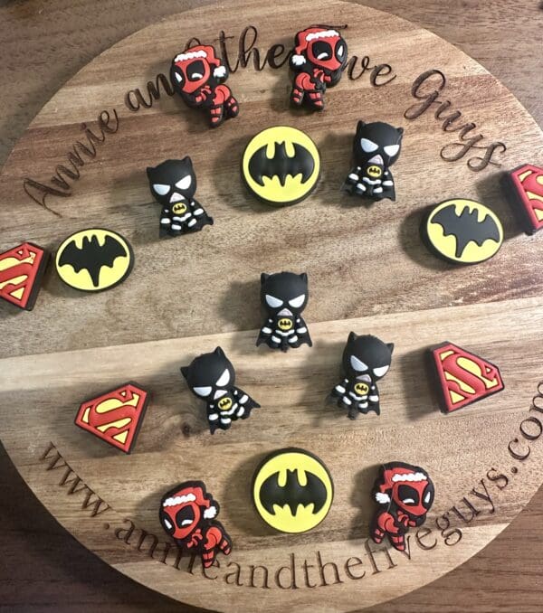 Round wooden board with toy figures of superheroes, featuring logos of Superman and Batman, arranged in a pattern. Website text on the board's edge.