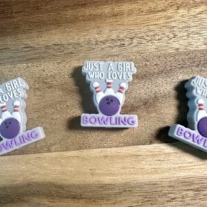 Pins with a purple bowling ball design, reading "Just a girl who loves bowling," placed on a wooden surface.