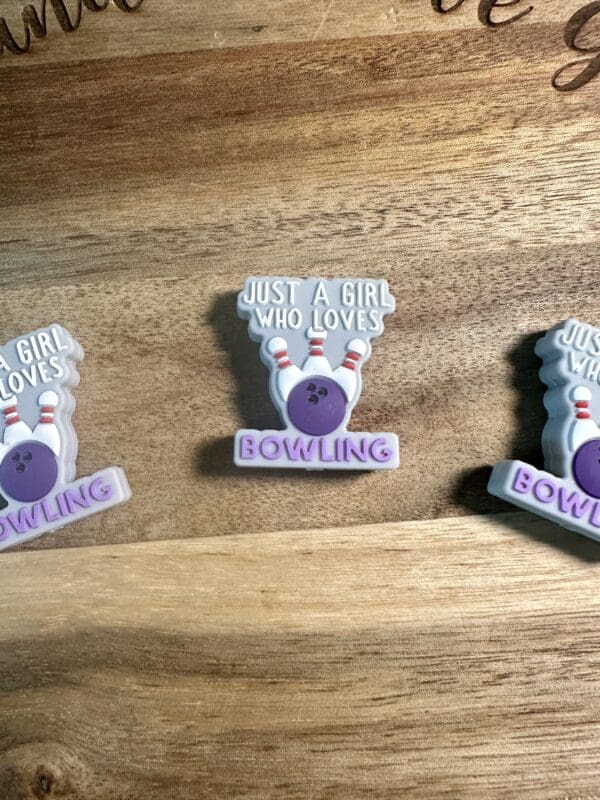 Pins with a purple bowling ball design, reading "Just a girl who loves bowling," placed on a wooden surface.