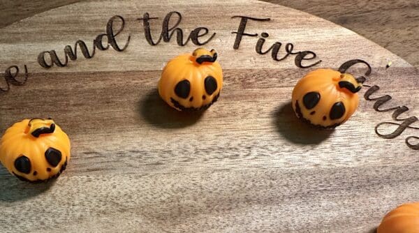 Three pumpkin-shaped orange chocolates with smiling faces on a wooden surface with partially visible lettering.