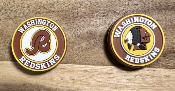 Two circular Washington Redskins logos on a wooden surface. One features a stylized "R" and the other shows a Native American profile.